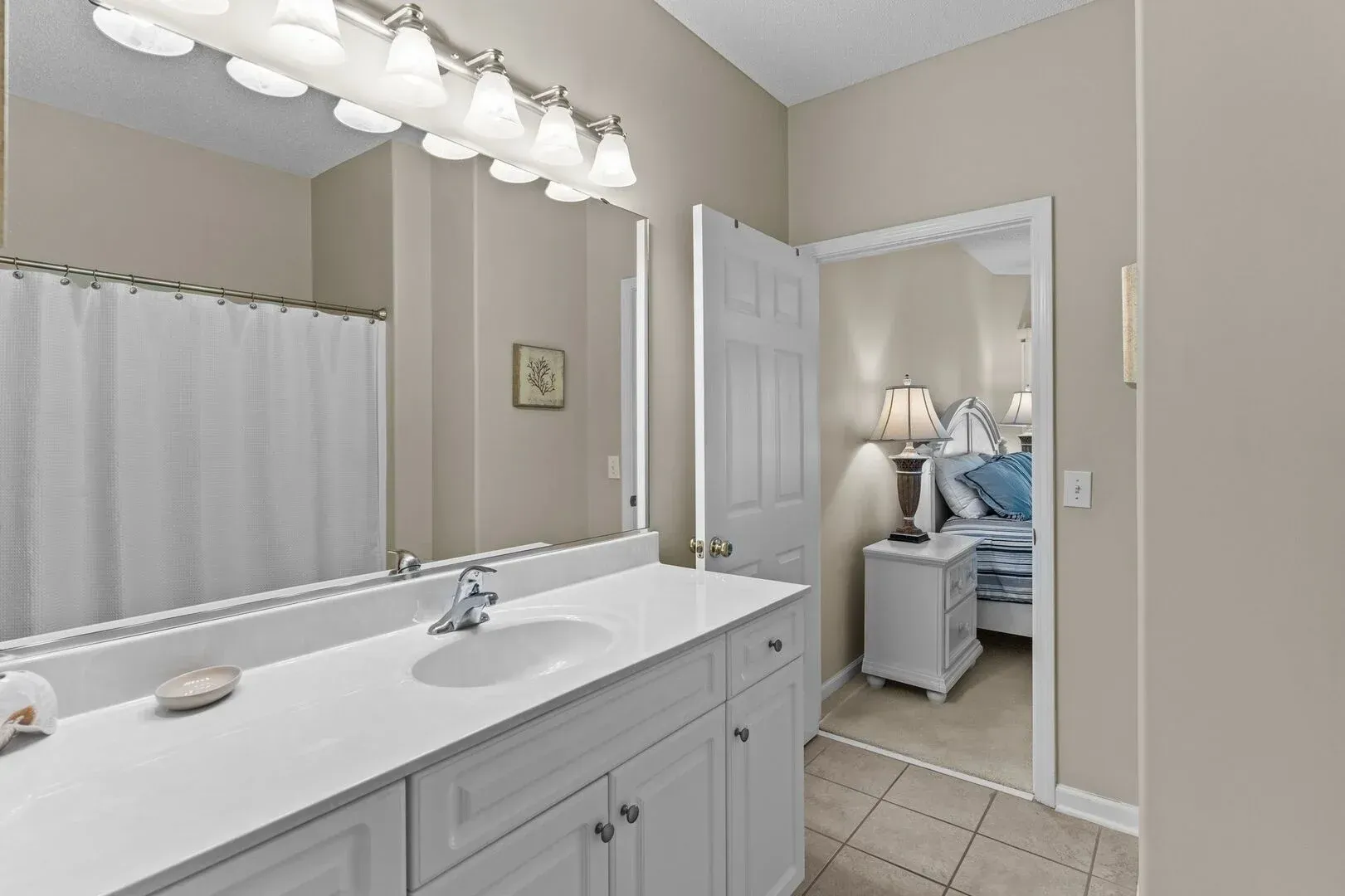 Bright bathroom vanity with mirror lights, white cabinets, and an open door to a bedroom with a blue bedspread