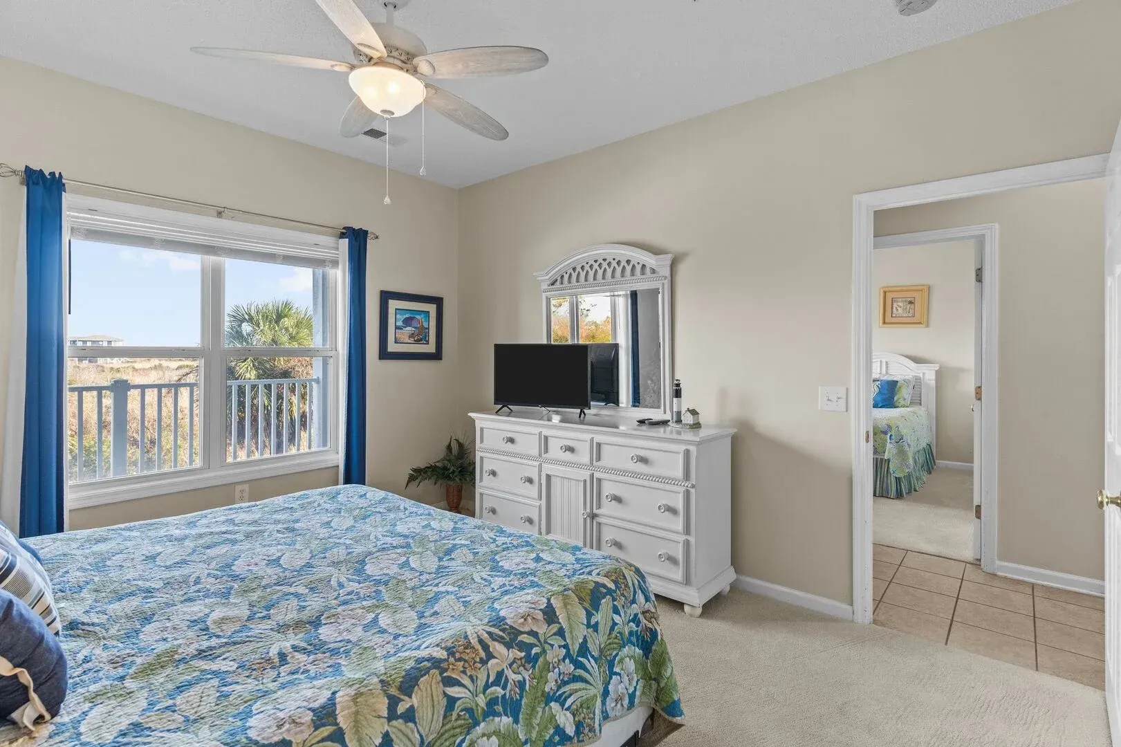 Bright bedroom with blue floral bedspread, white dresser, ceiling fan, and open bathroom doorway