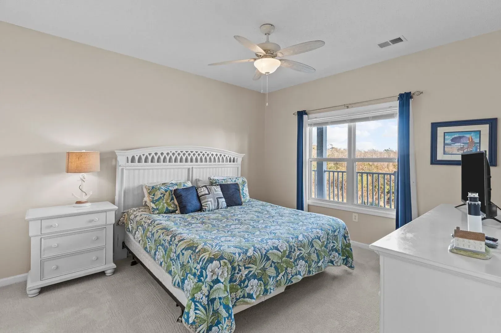 Bright bedroom with blue floral bedding, white furniture, and a window view of the water