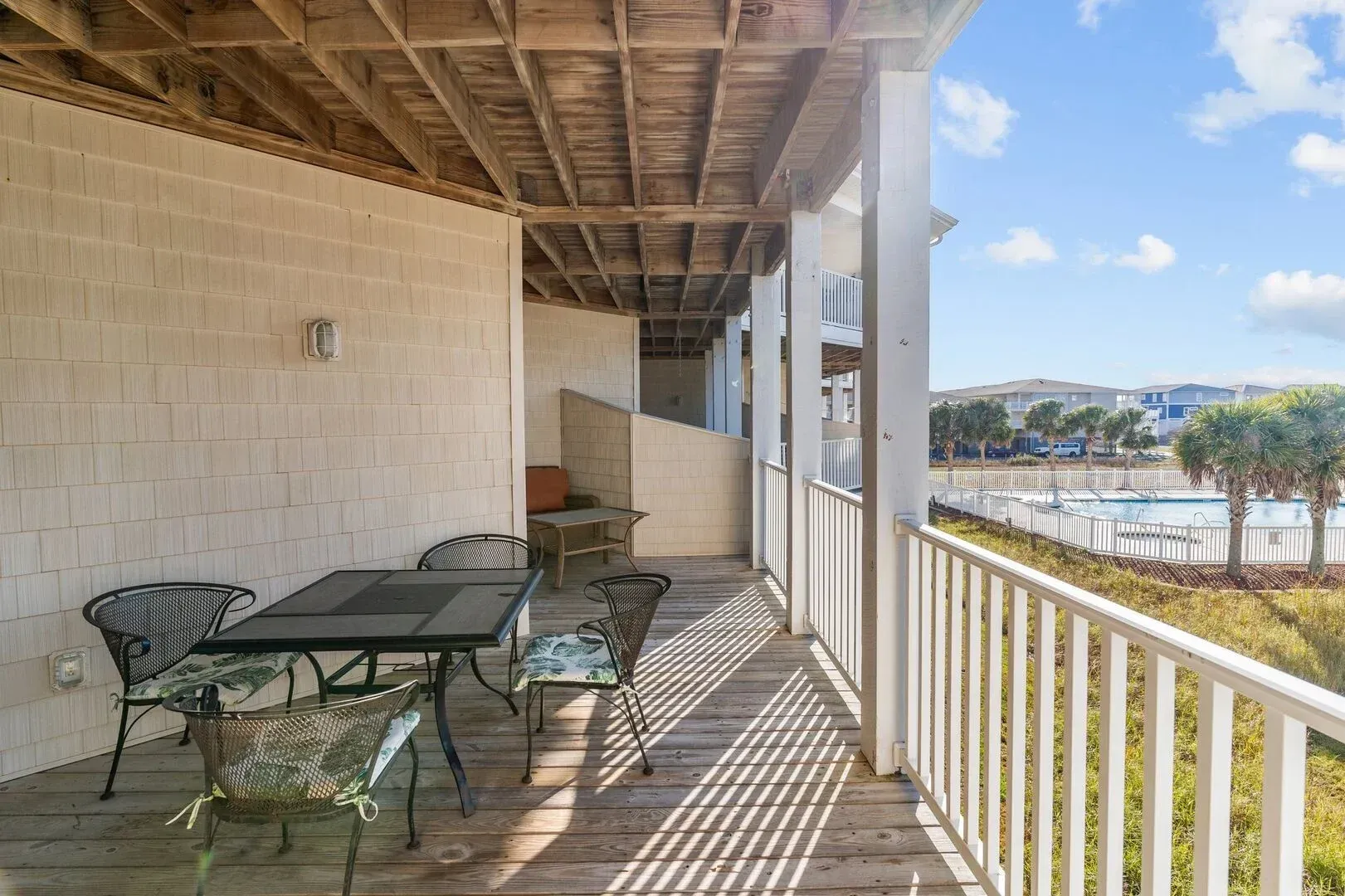 Covered balcony with glass table and chairs, overlooking a waterfront and blue sky.