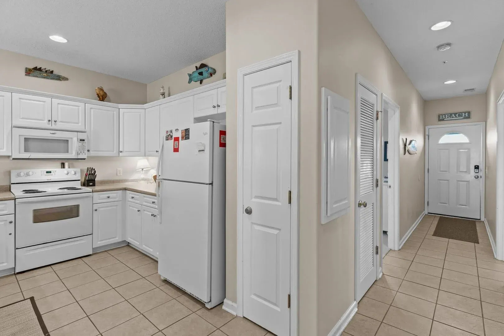 Bright white kitchen and hallway with tiled floor, white cabinets, and refrigerator on the left.