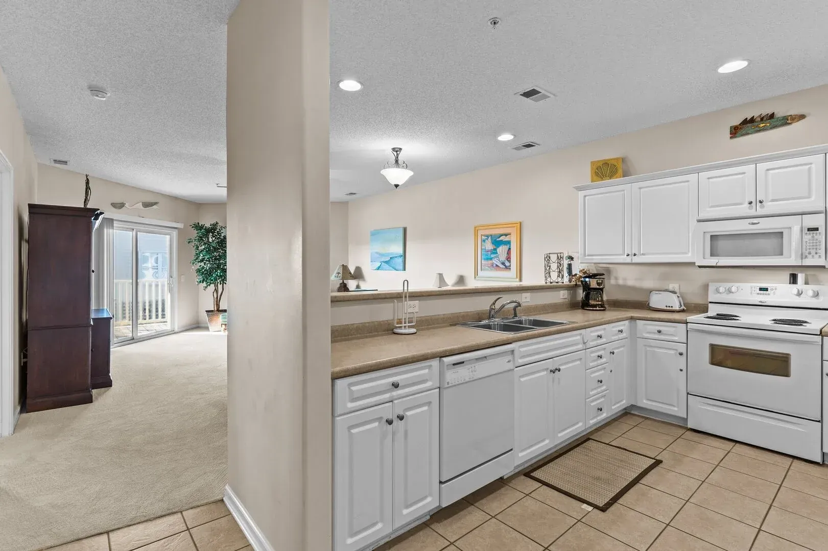Bright kitchen with white cabinets, tan countertops, tiled floor, and a view into a carpeted living area