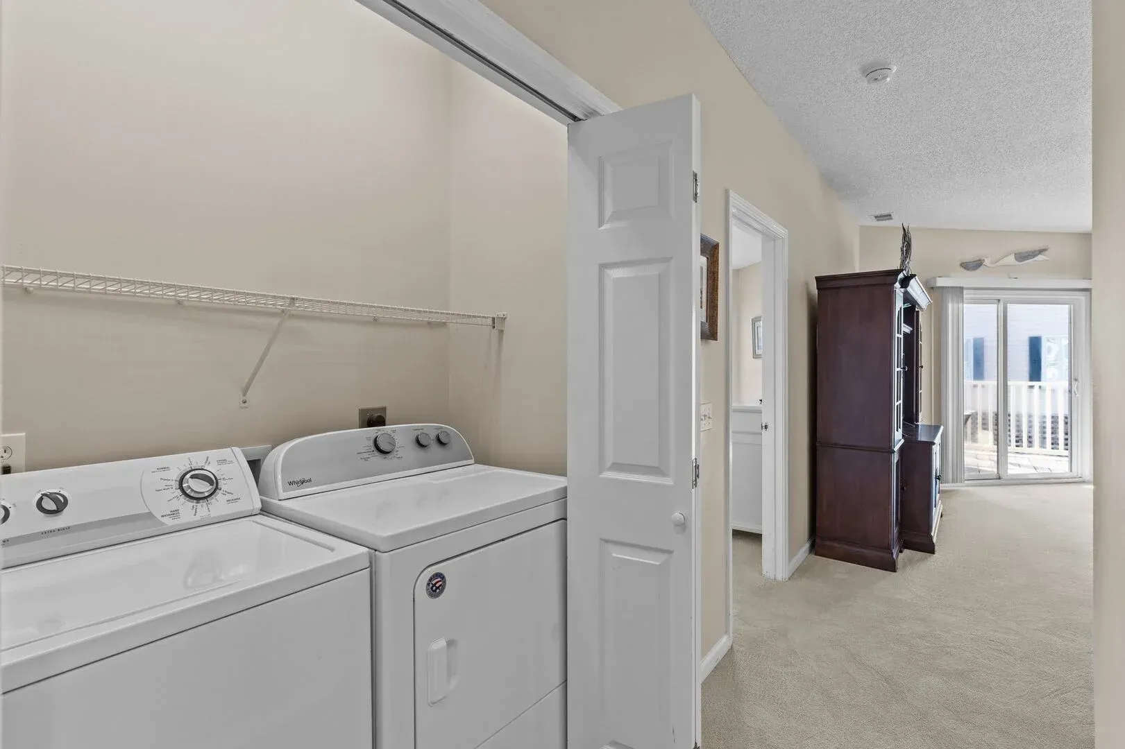 Laundry room with white washer and dryer beside a hallway leading to a bedroom and balcony.