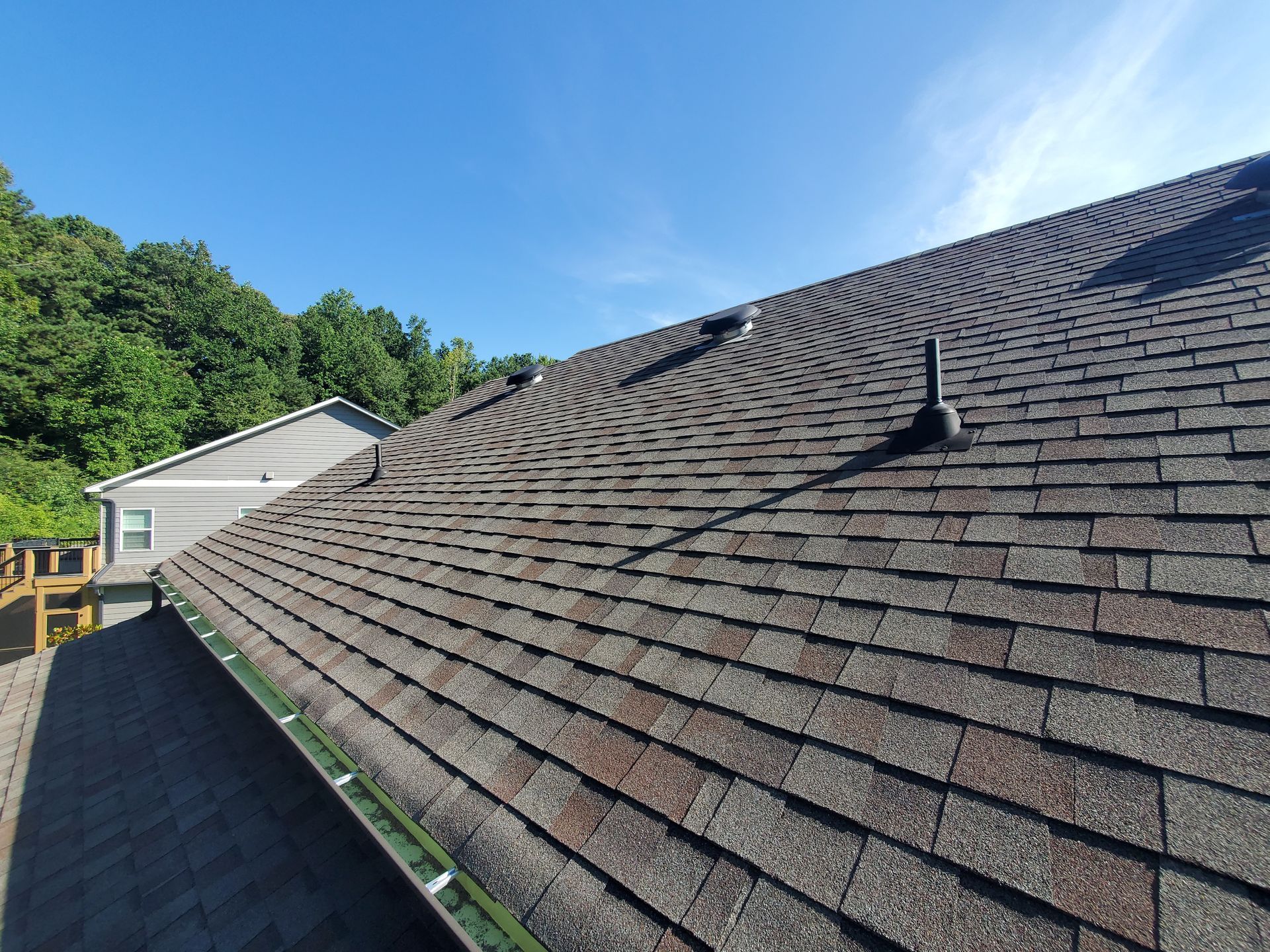 A brown shingle roof with vents, blue sky, and a treeline in the background.