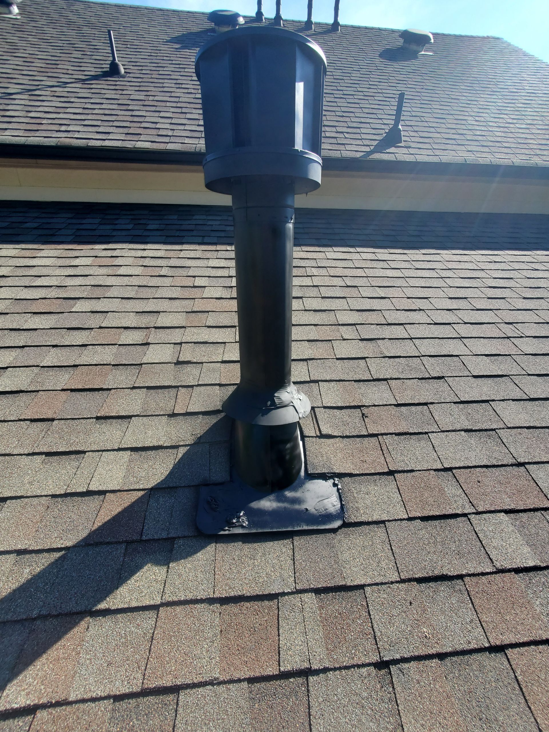 Black roof vent pipe on a shingle roof, casting a shadow in bright sunlight.