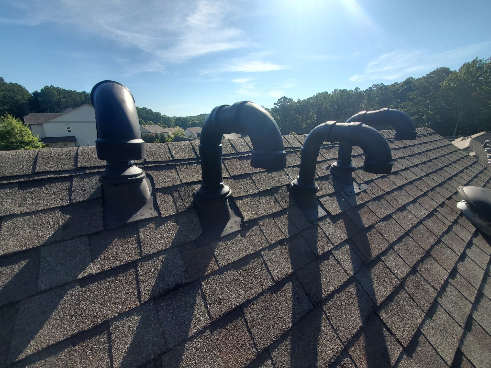 Black pipes on a shingled roof against a blue sky with trees in the background.