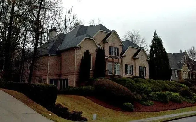 Brick house with black shutters, set on a grassy hill with landscaped shrubs and bare trees.