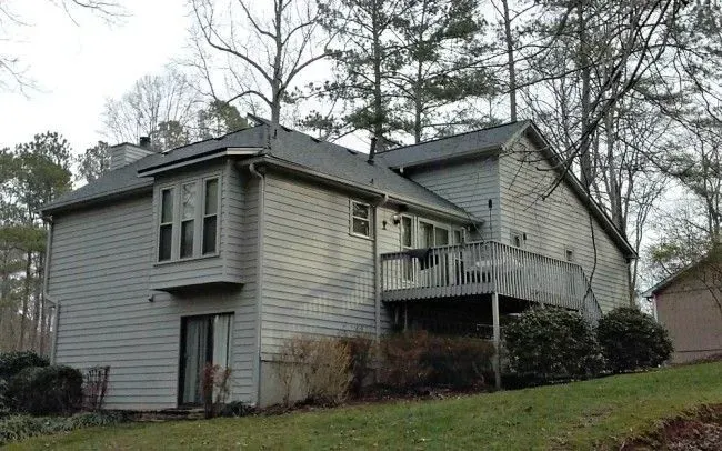Two-story gray house with a deck, surrounded by trees and bushes on a grassy hill.