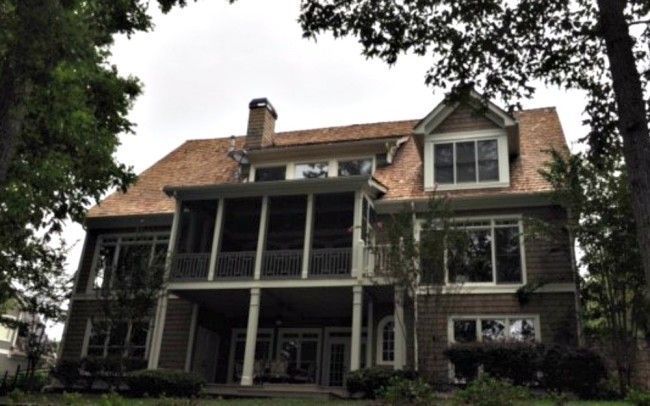 Back of a brown house with a screened porch, large windows, and a brown roof, viewed through trees.