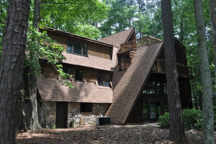 A-frame house in a forest setting with brown shingles, stone base, and wooden deck.