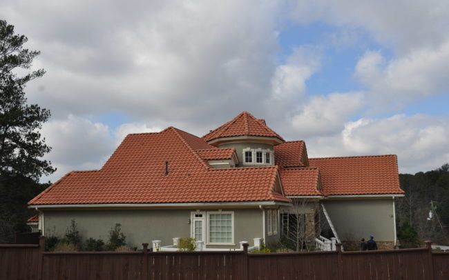 House with red tile roof and light green exterior behind a brown fence under a cloudy sky.
