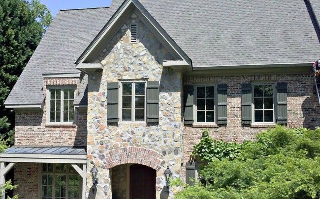 Stone and brick house with gray roof, green shutters, and arched entryway.