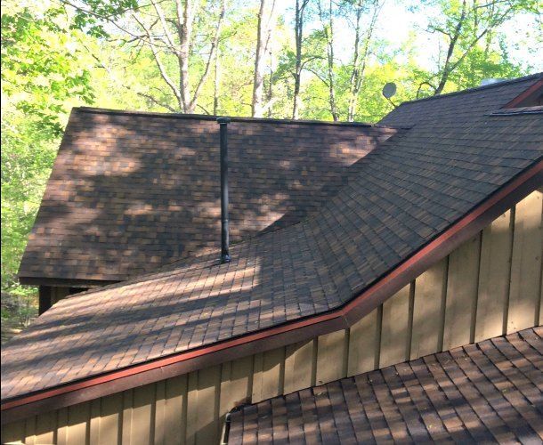 Brown shingled roof on a house, with a chimney. Surrounded by trees and a light brown wall.