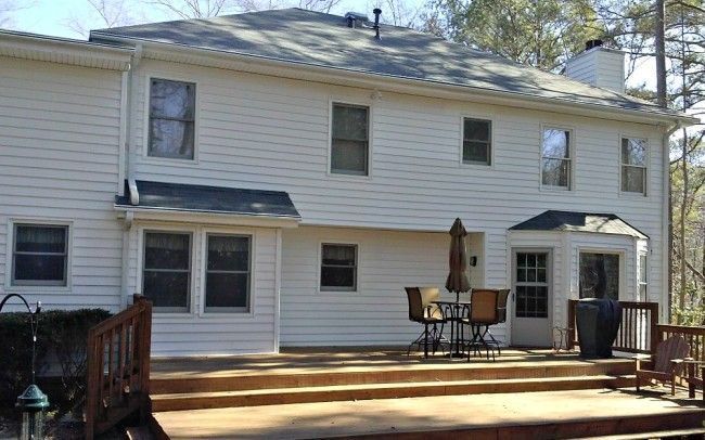 Back of a white two-story house with a wooden deck, patio furniture, and surrounding trees.