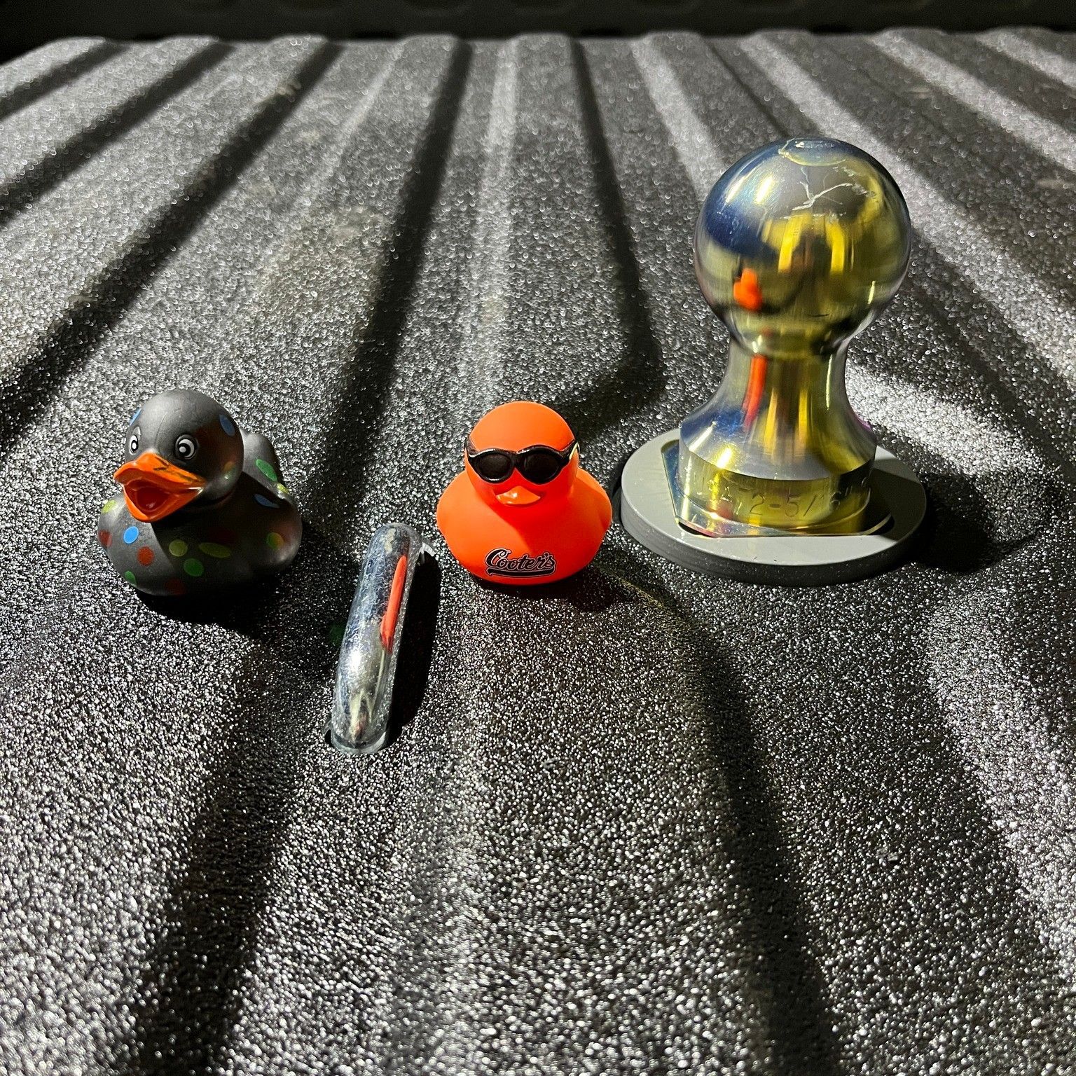 Two rubber ducks, a chrome bar, and a chrome hitch ball on a textured black surface.