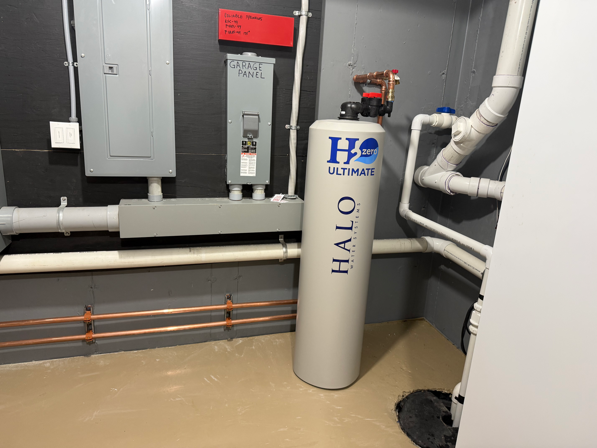 Utility room with gray walls, electrical panels, pipes, and a tall H2O filtration tank.