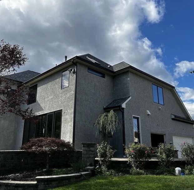 Gray stucco house with black framed windows, a dark roof, and cloudy sky.