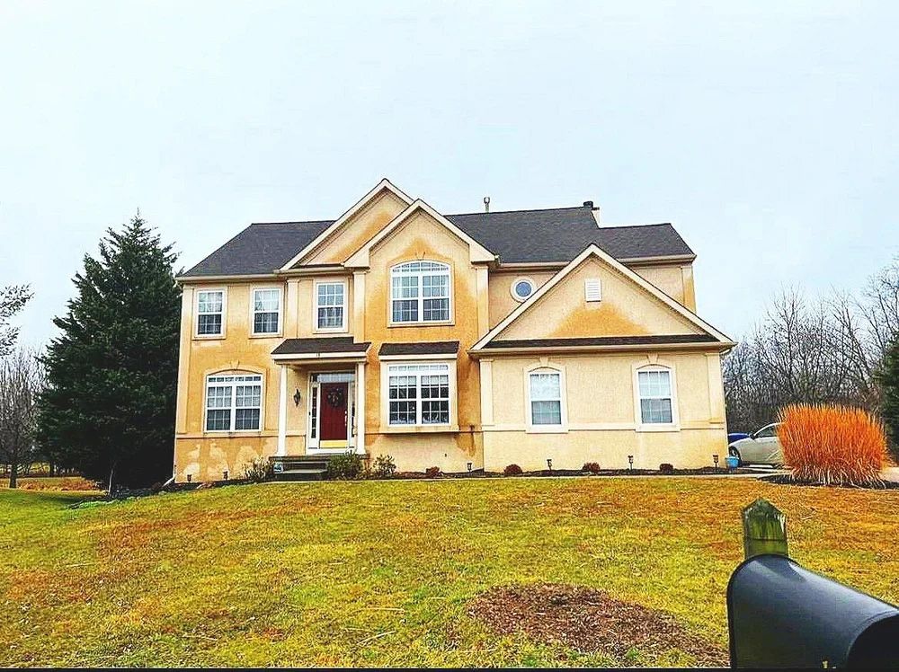 Yellow two-story house on a grassy hill; dark roof, red front door, mailbox in foreground, cloudy sky.