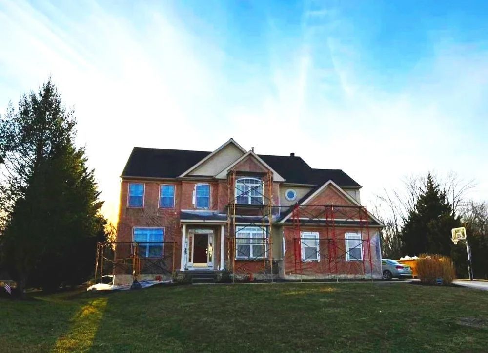 Two-story brick house with boarded windows, front lawn, and blue sky.