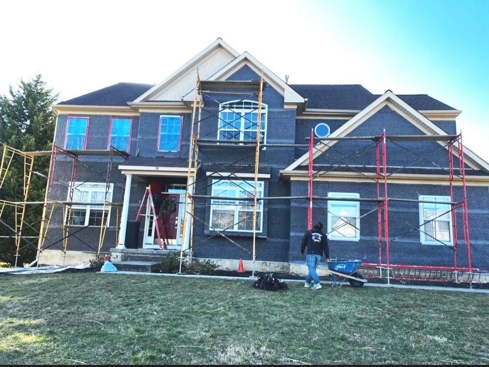 House exterior under construction, covered in dark wrap; scaffolding; worker with cart.