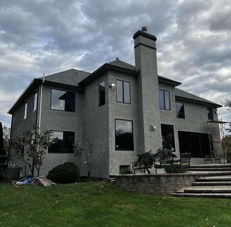 Gray stucco house with black-framed windows, tall chimney, and backyard steps under a cloudy sky.