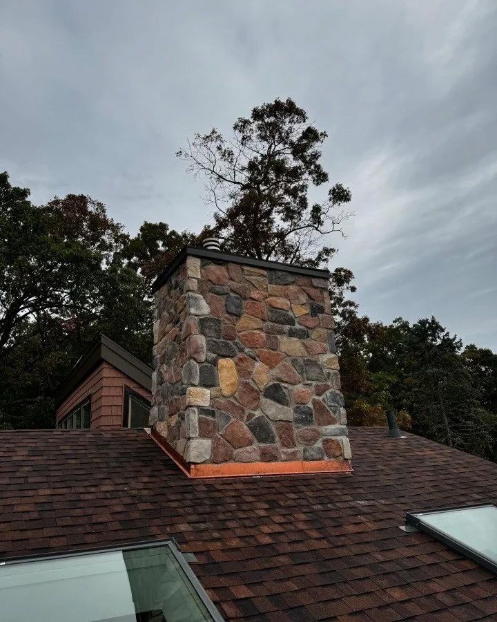 Stone chimney on a shingled roof with copper flashing, trees in background, cloudy sky.