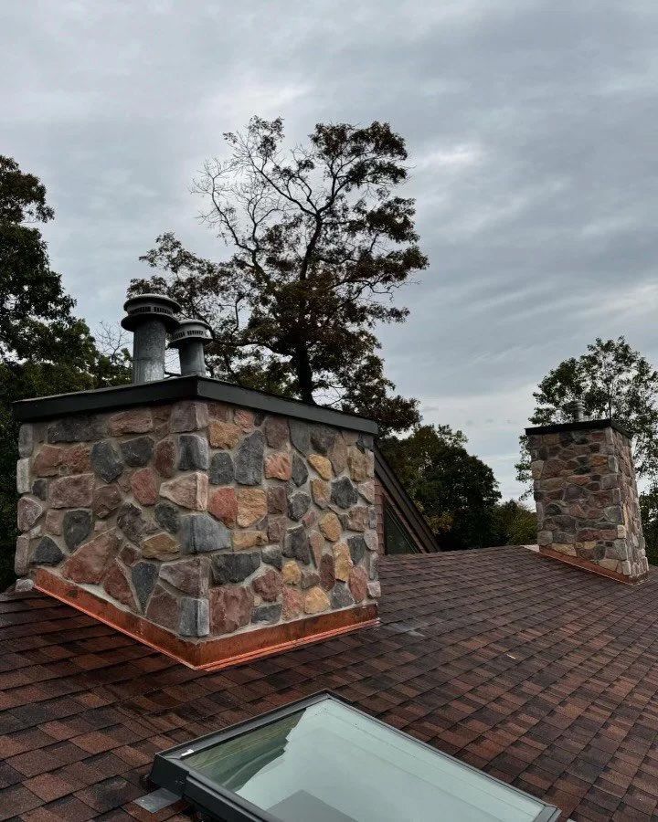 Stone chimneys with copper flashing on a shingle roof against a cloudy sky.