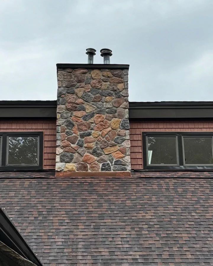 Stone chimney with two metal flues on a shingle roof. Red siding and windows flank the chimney.