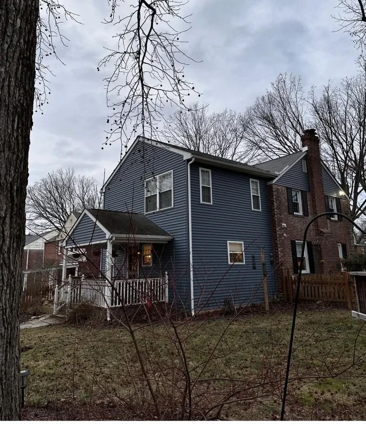 Two-story house with blue siding, brick chimney, porch, and bare trees under a cloudy sky.