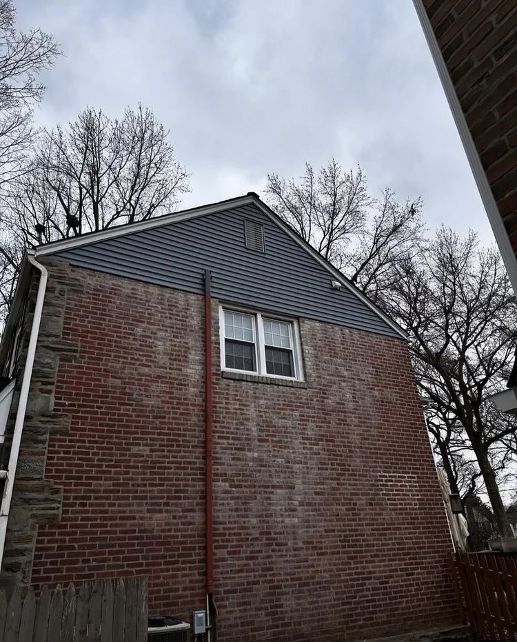 Brick house exterior with a gray roof and window; trees in the background.