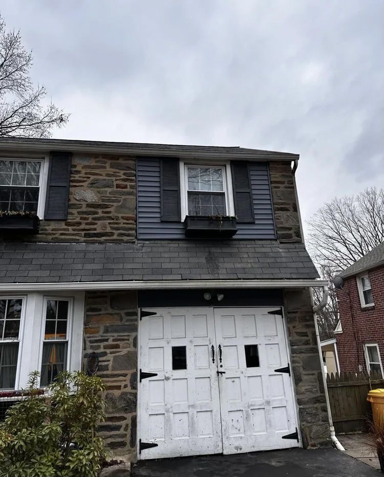 Stone house with a white garage door, dark shutters, and a gray roof under a cloudy sky.