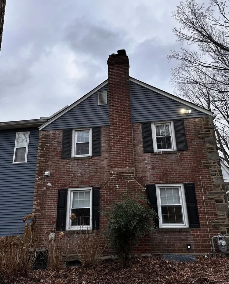 Brick house with a prominent chimney, dark shutters, blue siding, and a cloudy sky.