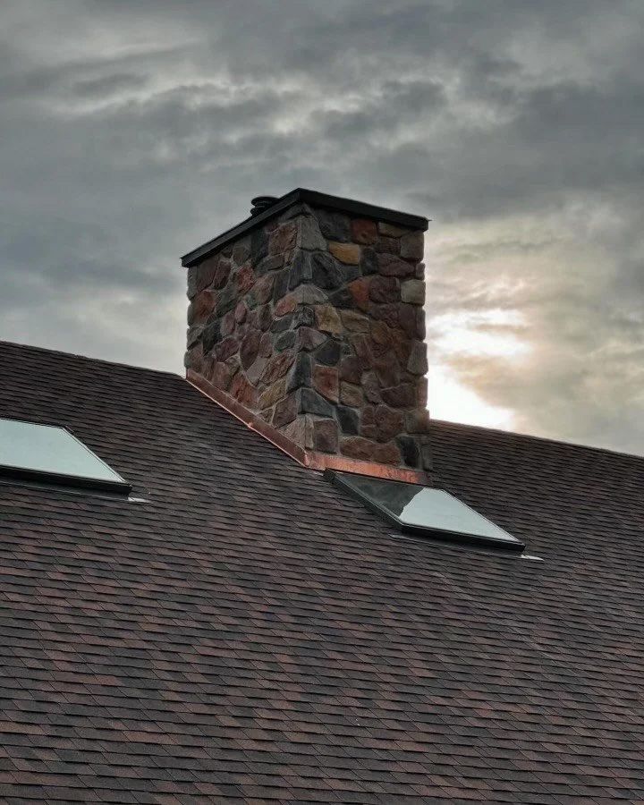 Stone chimney on a brown shingle roof against a cloudy sky.