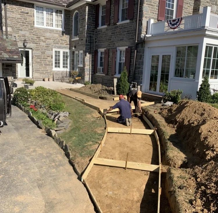 Construction workers building a concrete walkway next to a stone house; wooden forms are set.