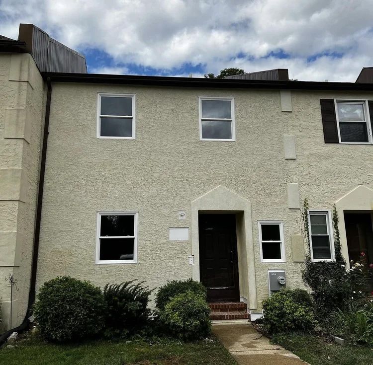 Two-story townhouse with light beige stucco facade, dark brown door, and white-framed windows. Bushes frame the entrance.