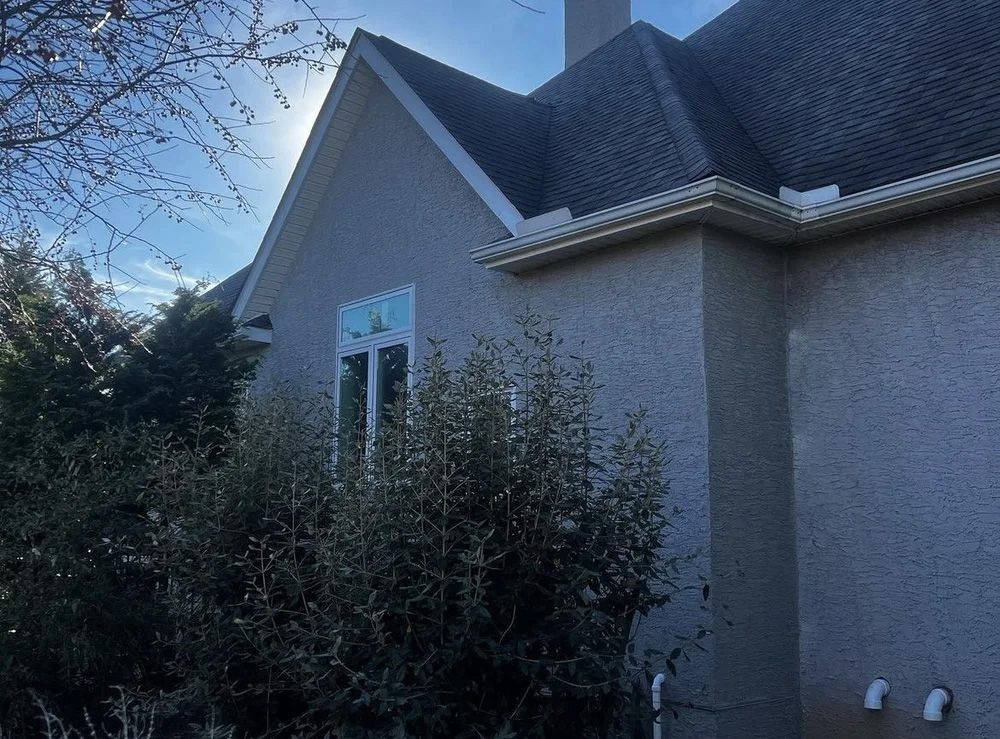 Side view of a house with stucco siding, dark roof, and a window. Sunlight shines through trees in front.