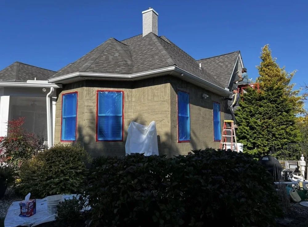House exterior being painted, windows taped with blue, ladder present, clear sky.