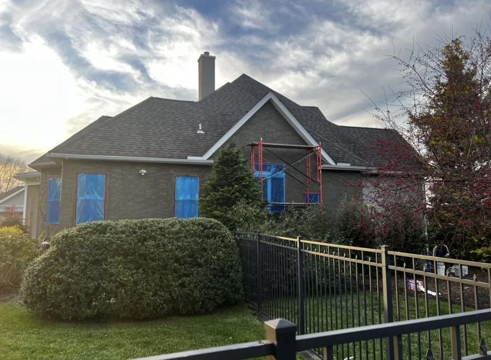 Brick house with boarded-up windows and scaffolding under a cloudy sky. A black fence is in the foreground.