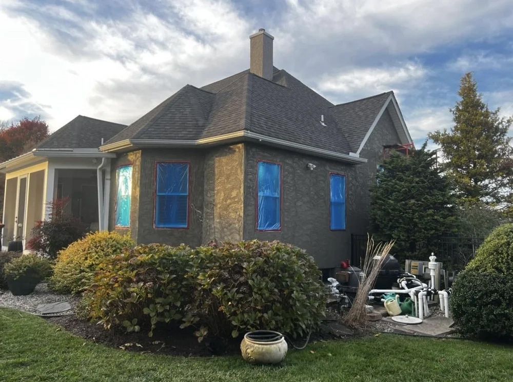 House with dark brown stucco exterior, blue-taped windows, and a dark shingled roof, set in a yard with bushes.