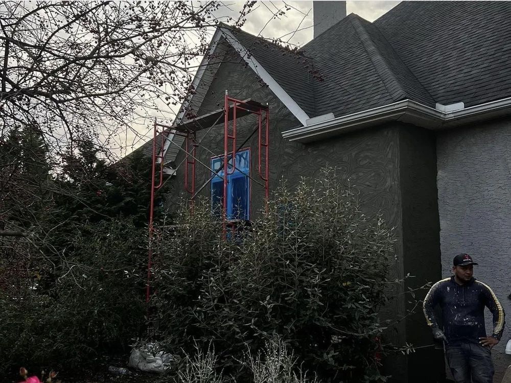 House exterior with scaffolding, bushes, and a worker. Grey stucco, cloudy sky.