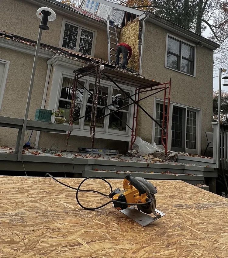 Construction worker on a roof, using a saw. Scaffolding, and plywood in the foreground. House exterior.