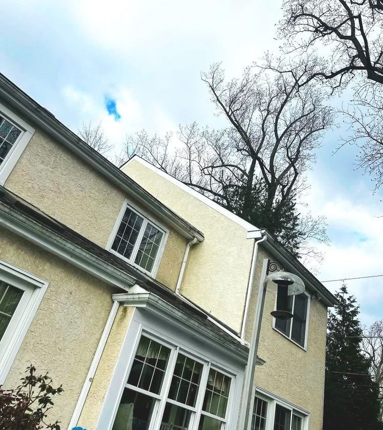 Two-story house with light yellow stucco siding, white trim, and a cloudy sky. Bare tree branches frame the top.