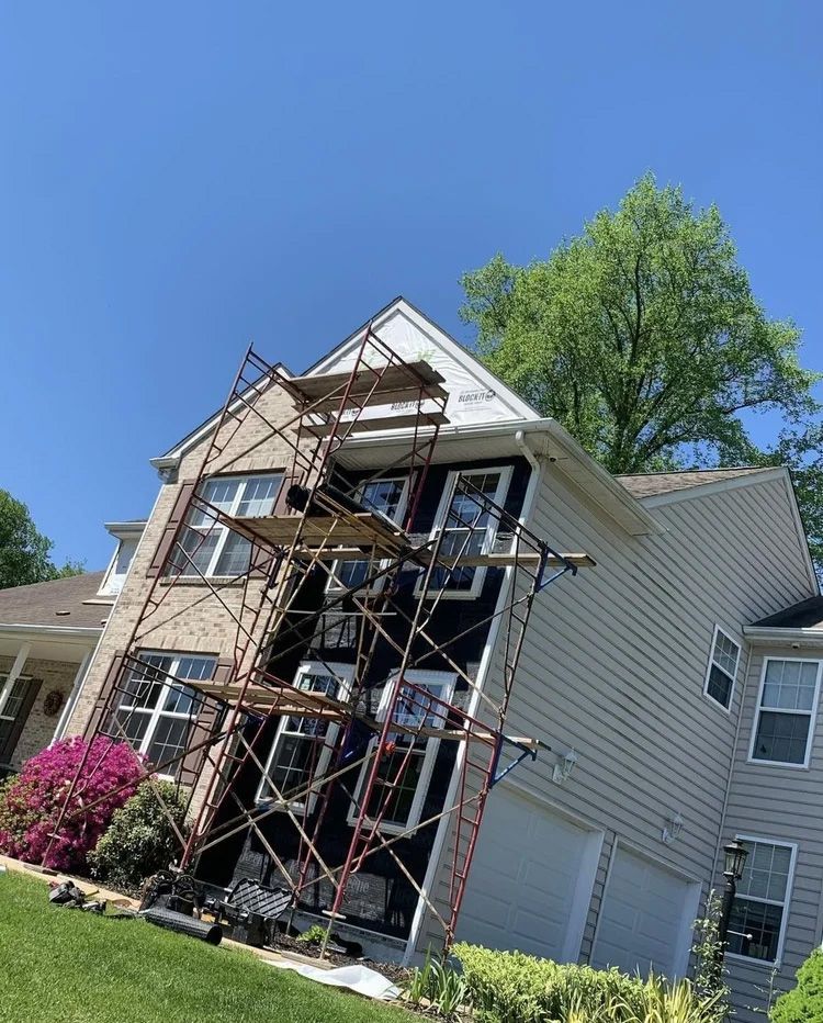 Scaffolding alongside a two-story house with black siding, beige siding, and a white roof on a sunny day.
