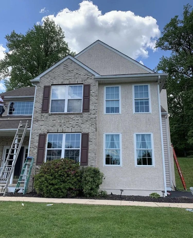 Two-story house with brick and stucco exterior, brown shutters, green lawn, ladder propped against side.