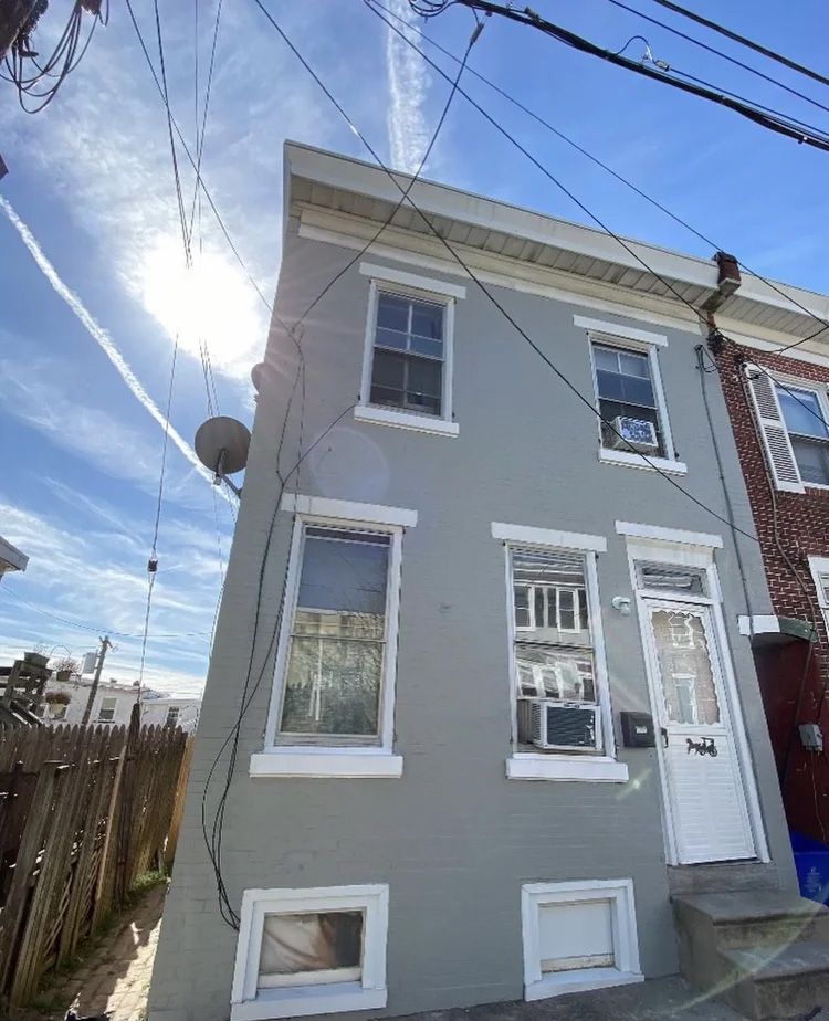 Gray two-story house with white trim and door. Power lines and satellite dish are visible. Bright sky.