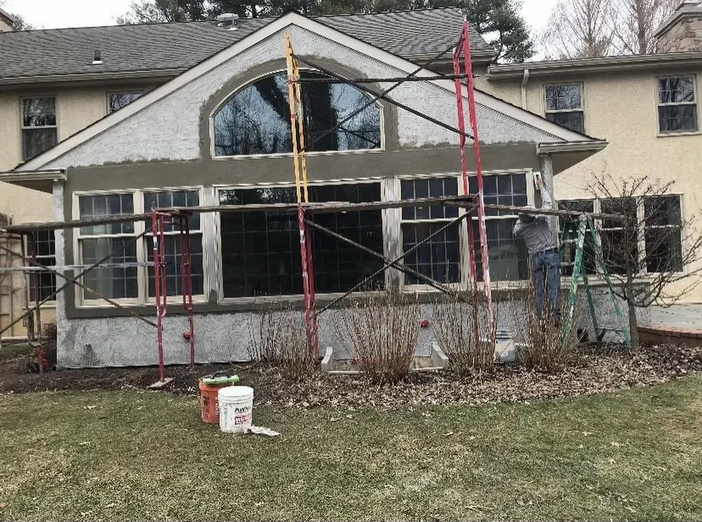 House with scaffolding; stucco exterior repair underway, front yard with grass and bushes.