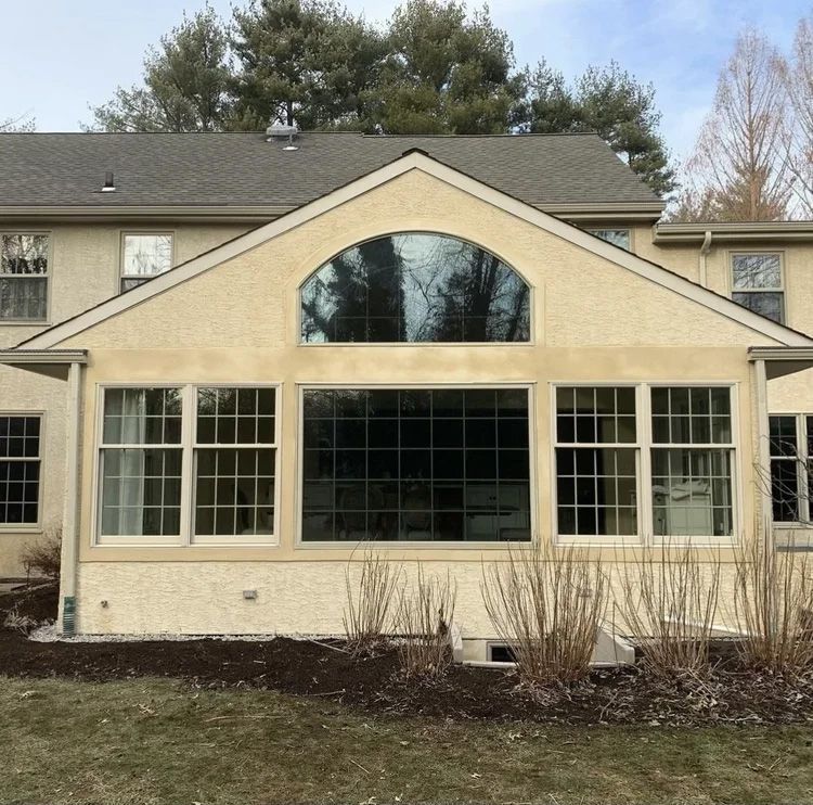 Tan house addition with arched window above a row of rectangular windows. Brown mulch, bare bushes, and grass.