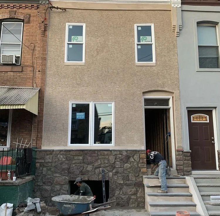 Workers renovating a two-story brownstone building. One mixes cement, another on stairs.