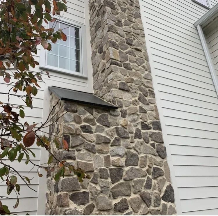 Exterior house with stone chimney and white siding, next to a window and tree branch.
