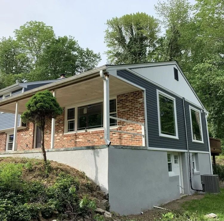 Two-story house with brick and blue siding, covered porch, and basement, nestled in a wooded area.